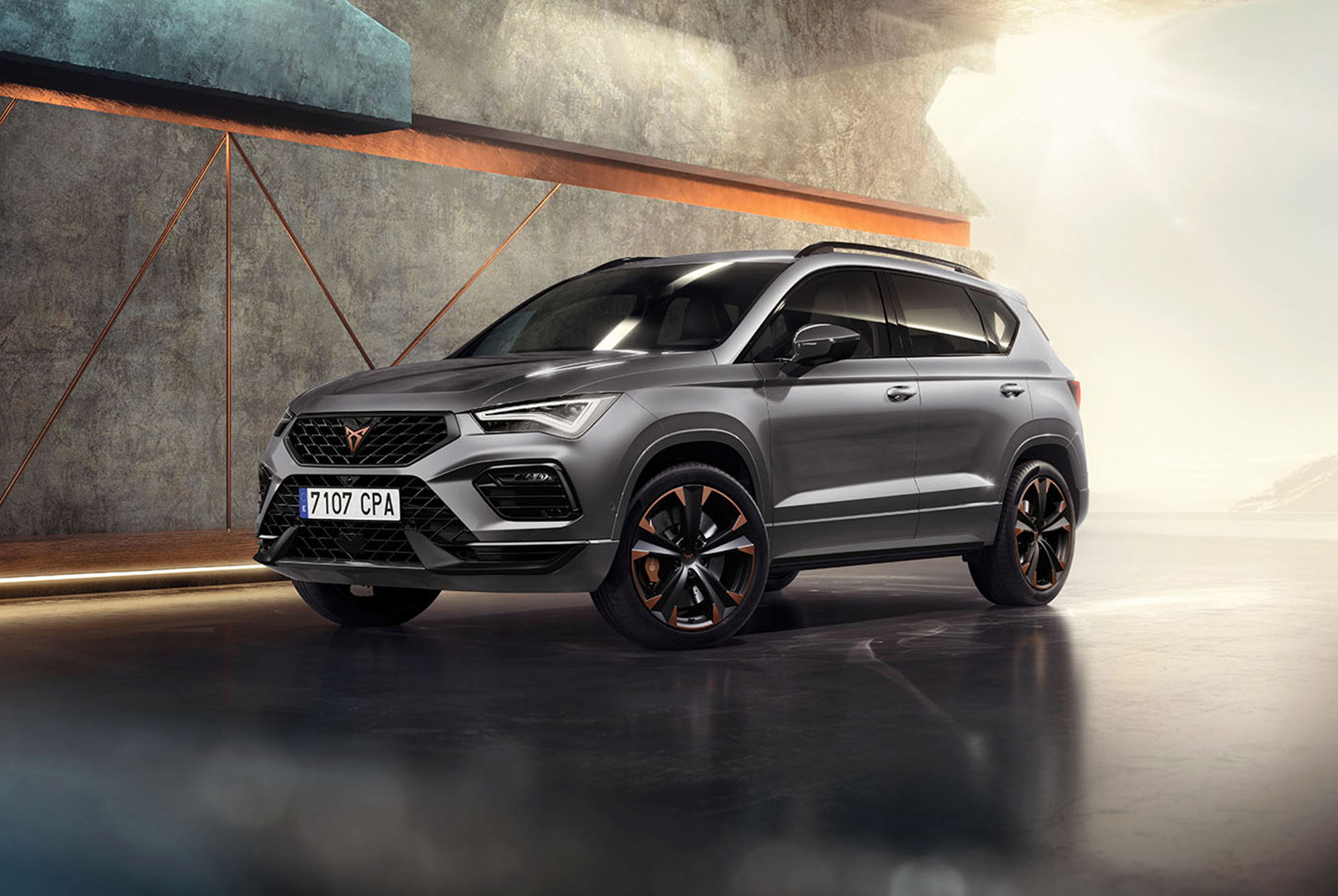  „Cupra Ateca“ – sportiškas ir kompaktiškas SUV, stovintis prie modernios betoninės sienos. Automobilio dizaine matomos aerodinamiškos linijos, LED priekiniai žibintai ir įspūdingi lengvojo lydinio ratlankiai su vario spalvos akcentais. Geometrinio dizaino grubi siena fone kontrastuoja su elegantišku automobilio dizainu.